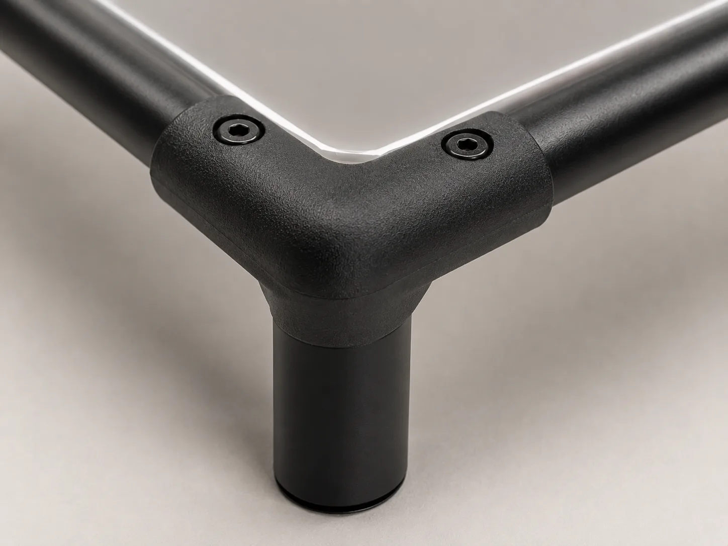 Extreme close-up of a matte black IronPaw elevated dog bed corner connector with two recessed hex screws, round black frame tubes, glossy grey PVC fabric, and a straight open-ended metal leg on a neutral studio background.