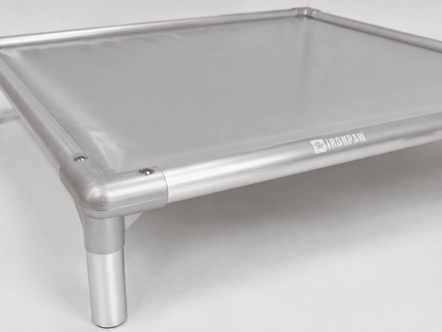Close-up of a grey PVC elevated dog bed corner with a brushed silver aluminium frame, rounded connector, flush top screws, bare tube leg, and pale IronPaw logo engraved on the front rail.