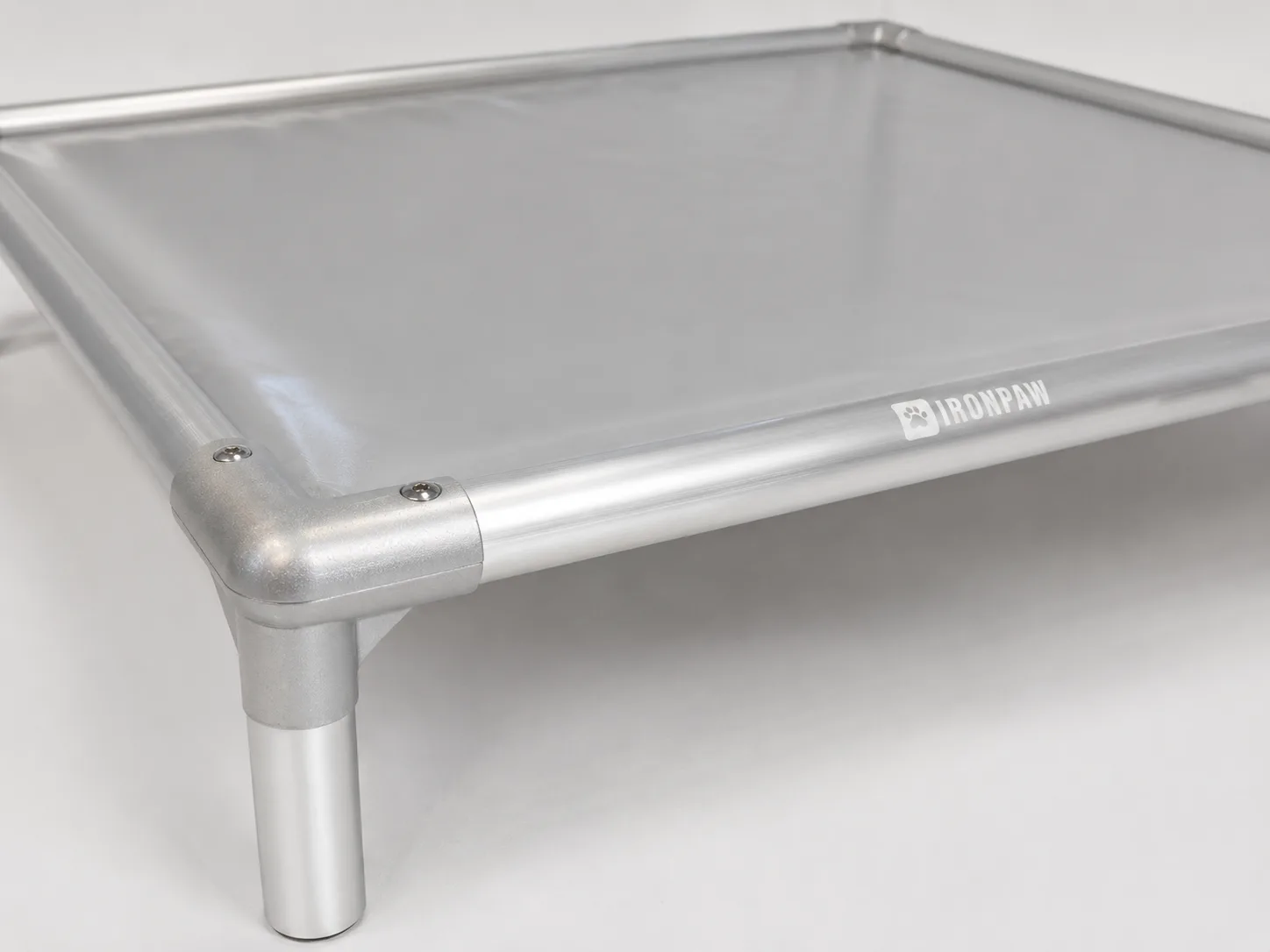Close-up of a grey PVC elevated dog bed corner with a brushed silver aluminium frame, rounded connector, flush top screws, bare tube leg, and pale IronPaw logo engraved on the front rail.